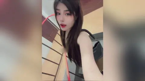 ThaiiTwix looks at the camera with long dark hair, wearing a black tank top and carrying a black handbag over her shoulder.