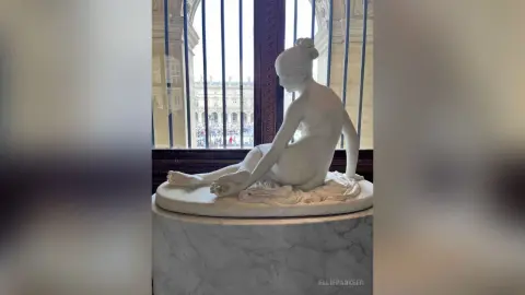 Elegant white marble sculpture of a seated nude woman displayed in a bright, classical museum setting.