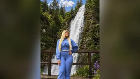 Blonde woman in a blue jumpsuit standing on a wooden bridge before a massive scenic waterfall in a lush forest.