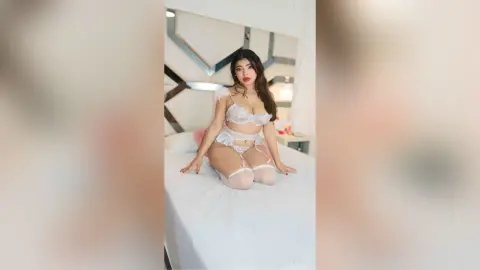 Young woman in white lace lingerie and stockings posing on a bed in a bright, modern bedroom.