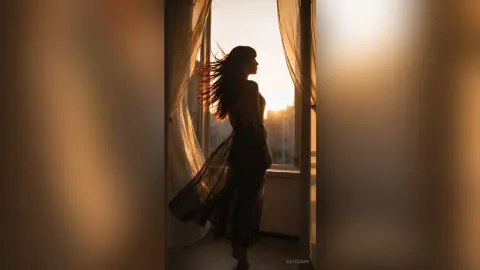 Silhouette of a woman in a flowing black dress standing by a window during a golden sunset mood.
