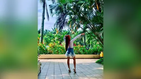 Young woman in a white top and denim skirt standing on a sunny patio surrounded by lush tropical palm trees.