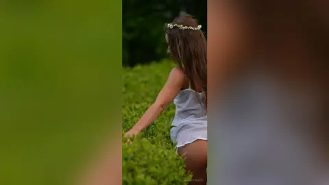 Dreamy woman in a white summer dress and flower crown walking through a lush green garden.