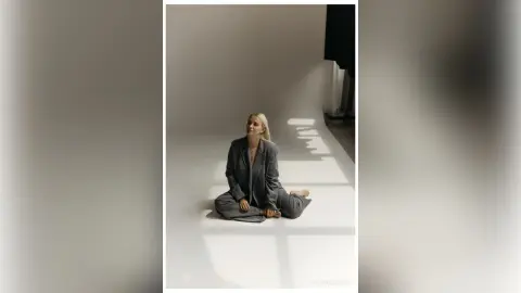 Elegant woman in a grey oversized suit sitting on a bright studio floor in a minimalist, serene setting.