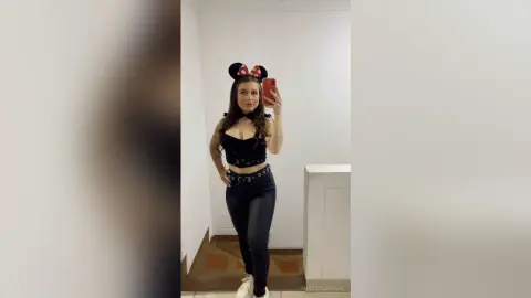 Young woman in a black crop top and dark jeans wearing Minnie Mouse ears for a playful indoor selfie.