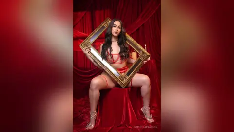 Glamorous woman in a red bikini posing with a gold frame against luxurious red velvet curtains.