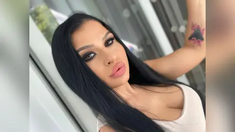 AlmaKane poses with long black hair and heavy eye makeup, wearing a white tank top while raising one arm to show a pink tattoo.