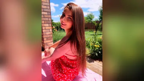 Smiling young woman in a red floral sundress sitting in a sunny garden on a bright summer day.
