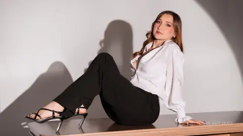 Elegant woman in white blouse and black trousers sitting on a platform against a minimalist studio background.