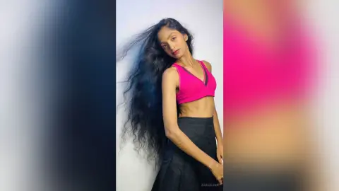 Young woman with long wavy hair wearing a pink crop top and black skirt posing against a white background.