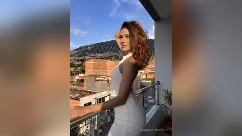 Young woman in a white dress standing on a balcony overlooking a city at sunset.
