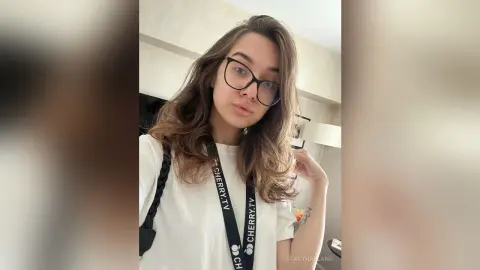StacyHarland wears black glasses and a white t-shirt while looking into the camera with her long brown hair down.