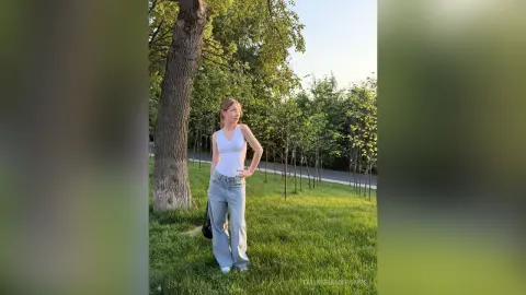 Young woman in a white tank top and light blue jeans standing in a sunny green park by a large tree.