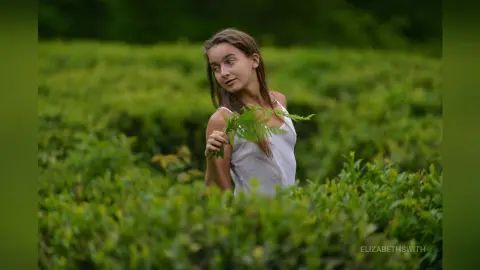 Young woman in a white tank top holding a green fern in a lush, dreamy tea plantation garden.