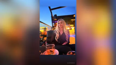 Blonde woman in a black shirt relaxing at a rooftop lounge bar during a sunset evening.