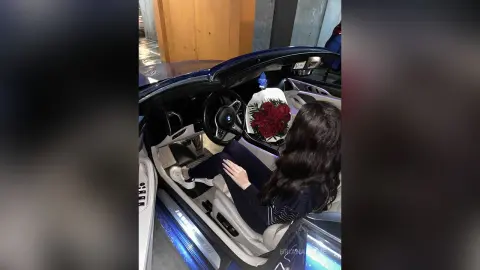 Woman in dark clothing sitting in a luxury convertible car with red roses on the passenger seat.
