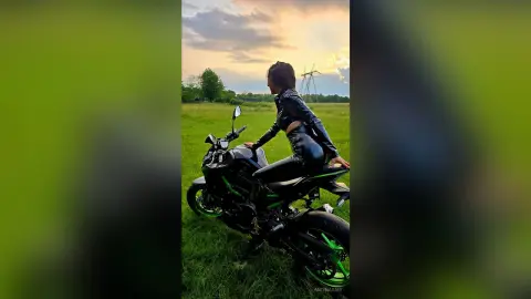 Stylish woman in black leather outfit riding a black motorcycle through a green field at sunset.
