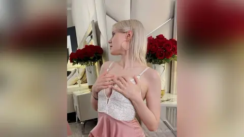 Blonde woman in a white lace camisole and pink satin skirt posing elegantly near red roses in a bright room.