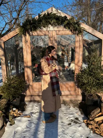 Woman in a beige coat and plaid scarf standing by a wooden greenhouse in a snowy winter garden.