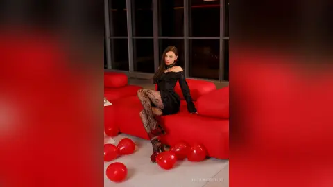Elegant woman in a black cocktail dress sitting on a red sofa surrounded by red heart balloons in a moody room.
