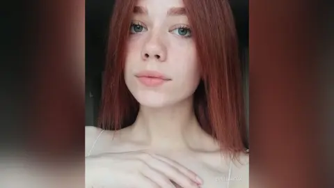 Portrait of a young woman with red hair and freckles wearing a white strap top in soft natural lighting.
