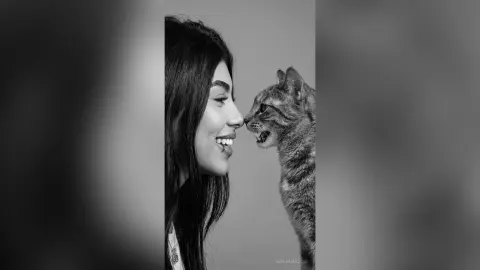 Black and white portrait of a smiling young woman sharing a tender moment with a tabby cat in a minimalist setting.