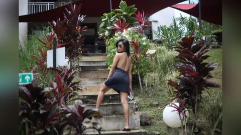 MokaAkashiya — Young woman in a black mini dress posing on wooden stairs amidst lush tropical plants in a serene garden setting.