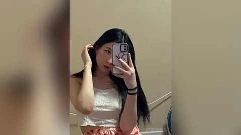 Young woman with long dark hair in a white crop top taking a moody mirror selfie in a minimalist room.