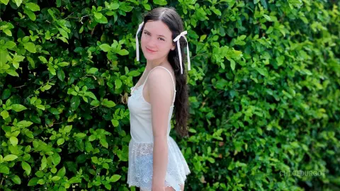 Smiling young woman in a white lace summer dress standing before a lush green garden hedge.