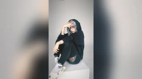 Trendy young woman in a black hoodie and dark jeans sitting on a white platform in a minimalist studio setting.