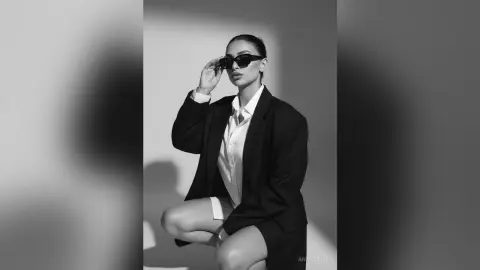 Stylish woman in a black blazer and white shirt wearing sunglasses in a moody studio setting.