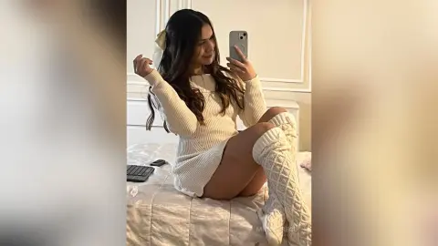 Young woman in a cozy white knit sweater dress and matching leg warmers sitting on a bed in a bright room.
