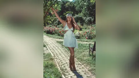 AlessiaCrush poses in a white mini dress and brown boots on a garden path, reaching up toward a tree branch.