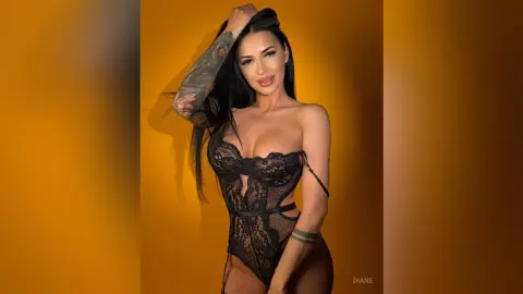 Smiling tattooed woman in a black lace bodysuit posing against a warm yellow background.