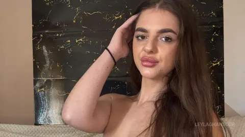 Young woman with long brown hair posing in a bedroom setting with a dark marble headboard.