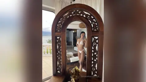 Young woman in a white bikini taking a mirror selfie in a bright, tropical resort room.