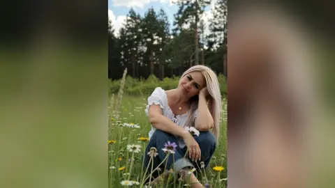 Blonde woman in a white ruffled top sitting peacefully in a sunny wildflower meadow near a pine forest.