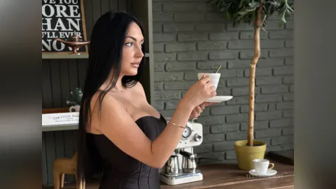 TessaWinsent — TessaWinsent in a black strapless top holds a white coffee cup in a cafe setting.