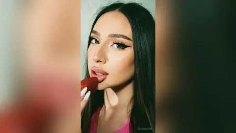 Young woman with winged eyeliner and long dark hair eating a strawberry in a soft, warm indoor setting.