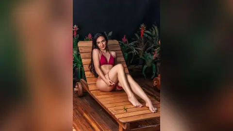 Smiling woman in a red bikini relaxing on a wooden lounge chair in a lush tropical garden setting.