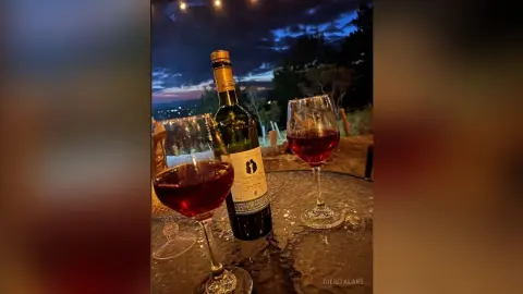 Red wine in glasses next to a bottle on a glass table during a romantic outdoor sunset dinner.