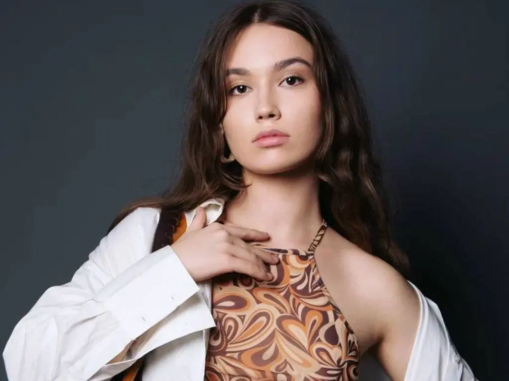 Young woman in a retro swirl print halter top and white shirt posing with a moody expression against a dark backdrop.