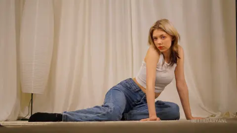 Young woman in white tank top and blue jeans sitting on floor against cream curtains in soft lighting.