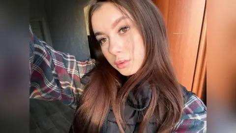 Young woman with long brown hair wearing a plaid shirt and black puffer vest in warm indoor lighting.