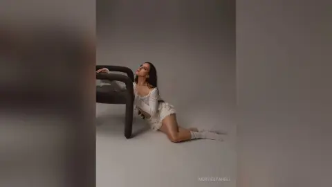Elegant woman in a white lace outfit sitting on the floor by a dark chair in a minimalist studio setting.
