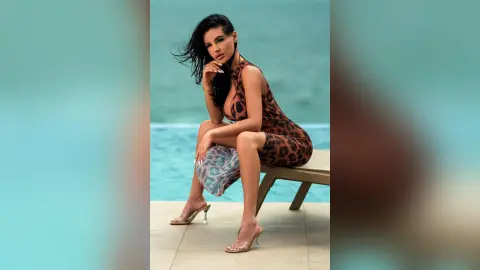 Elegant woman in a leopard print maxi dress sitting by the ocean in a glamorous summer setting.