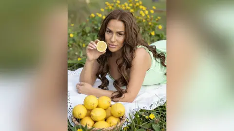 Kayla lies on a white blanket in a field of yellow flowers, wearing a light green top and holding a lemon slice near her mouth.