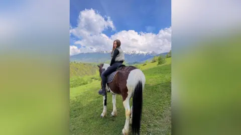 EvelynMills sits upright on a brown and white horse in a grassy field with mountains in the background.