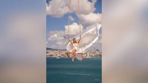 Anita sits on a wooden swing wearing a white dress against a blue sky with white clouds.