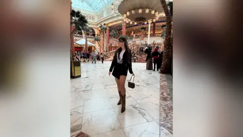 Stylish woman in a black outfit and boots walking through a luxurious, sunlit marble shopping mall lobby.